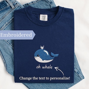 May include: Navy blue t-shirt with an embroidered blue whale design and the text "oh whale". The shirt is folded on top of a pair of blue jeans. The text "Change the text to personalize!" is also visible.