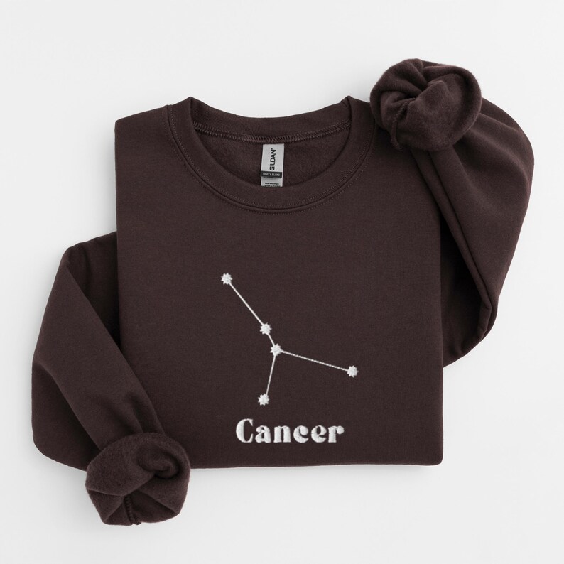 Peut inclure: Un sweat-shirt marron fonc&eacute; avec une constellation du zodiaque Cancer brod&eacute;e en blanc et le mot "Cancer" en dessous.