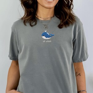 May include: A gray t-shirt featuring a blue embroidered whale design with the text "oh whale" in a script font. The whale has a small crown. The shirt has short sleeves and a relaxed fit.
