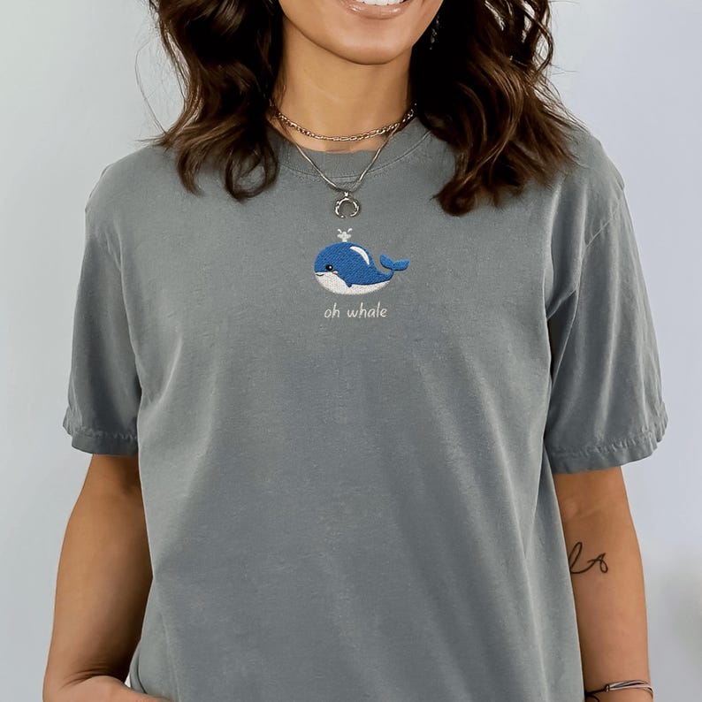 May include: A gray t-shirt featuring a blue embroidered whale design with the text "oh whale" in a script font. The whale has a small crown. The shirt has short sleeves and a relaxed fit.
