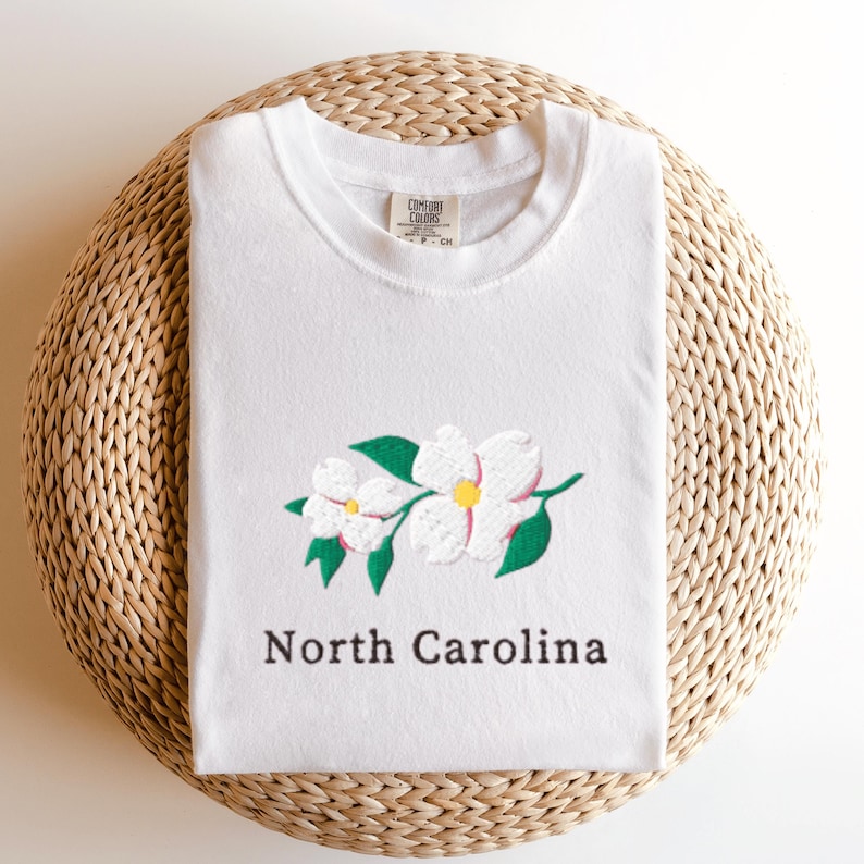 May include: A white short-sleeve t-shirt featuring an embroidered graphic of two white dogwood flowers with green leaves and yellow centers. The text North Carolina is printed in black below the flowers. The shirt has a Comfort Colors brand label.
