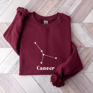 Peut inclure: Sweat-shirt bordeaux avec une constellation du zodiaque Cancer brod&eacute;e en blanc et le mot "Cancer" en dessous.