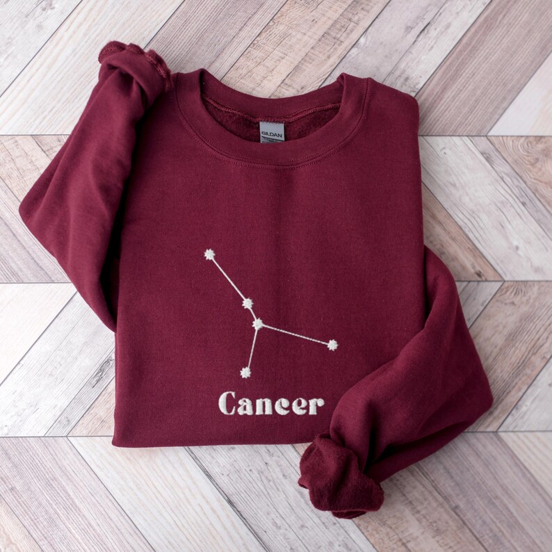 Peut inclure: Sweat-shirt bordeaux avec une constellation du zodiaque Cancer brod&eacute;e en blanc et le mot "Cancer" en dessous.