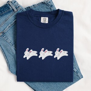 May include: Navy blue t-shirt with three embroidered white rabbits with pink ears. The shirt is displayed on top of a pair of blue denim jeans. The shirt has a Comfort Colors tag.