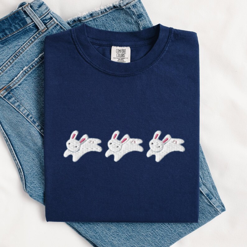 May include: Navy blue t-shirt with three embroidered white rabbits with pink ears. The shirt is displayed on top of a pair of blue denim jeans. The shirt has a Comfort Colors tag.