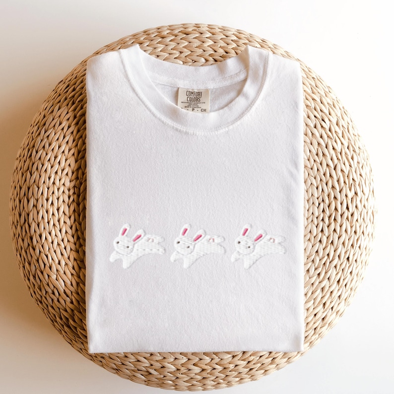 May include: White t-shirt with a crew neck featuring three embroidered white rabbits with pink ears. The shirt is made of a soft material and is displayed on a woven, circular surface. The tag reads "Comfort Colors".