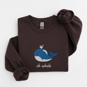 May include: A dark brown sweatshirt with a blue whale embroidered on the front. The whale has a small white spout and a white belly. The text "oh whale" is embroidered below the whale.