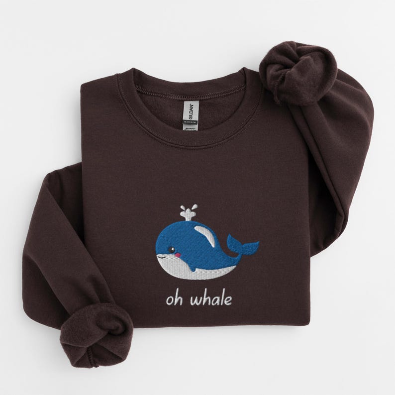 May include: A dark brown sweatshirt with a blue whale embroidered on the front. The whale has a small white spout and a white belly. The text "oh whale" is embroidered below the whale.