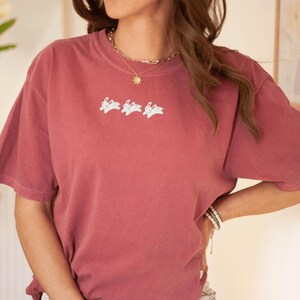 May include: A dusty rose-colored t-shirt with a crew neck and short sleeves. The shirt features a white embroidered design of three rabbits. The shirt is made of a soft, comfortable material.