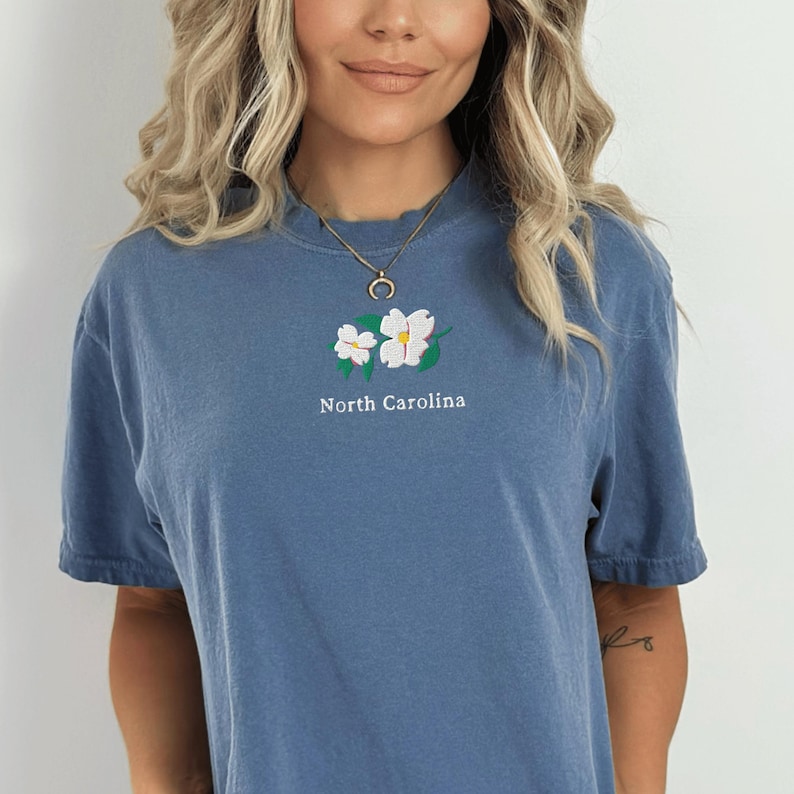 May include: A blue short-sleeve t-shirt featuring an embroidered design of two white dogwood flowers with green leaves centered on the chest. Below the floral embroidery, the text North Carolina is printed in a simple white sans-serif font.