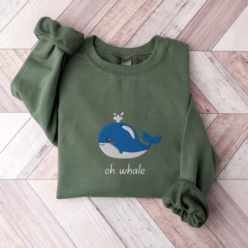 May include: A green sweatshirt with a blue whale embroidered on the front. The whale has a white spout and a white belly. The text "oh whale" is embroidered below the whale.