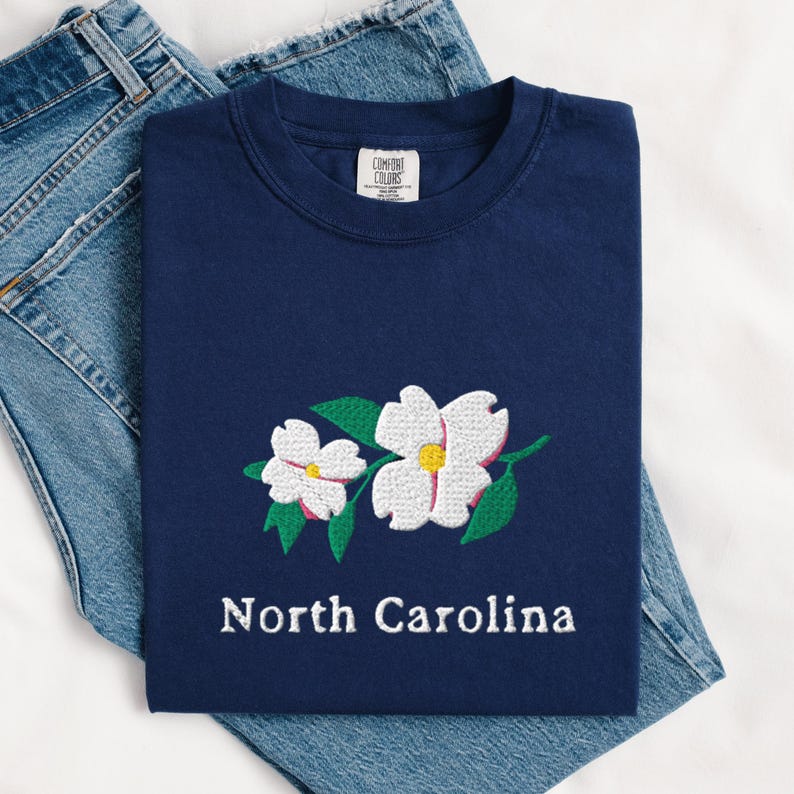 May include: A navy blue t-shirt featuring an embroidered design of two white dogwood flowers with yellow centers and green leaves. Below the floral embroidery, the text North Carolina is stitched in white. The shirt has a Comfort Colors label.