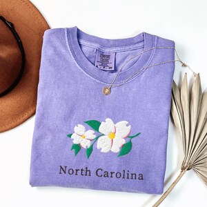 May include: A lavender short-sleeved t-shirt featuring an embroidered graphic of two white dogwood flowers with yellow centers and green leaves. The text North Carolina is printed below the flowers. The shirt has a Comfort Colors brand neck label.