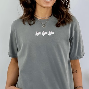 May include: A gray t-shirt featuring three white embroidered rabbits with pink accents. The shirt has short sleeves and a relaxed fit. The rabbits are in a running pose, adding a playful touch to the casual top.