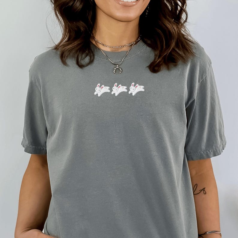 May include: A gray t-shirt featuring three white embroidered rabbits with pink accents. The shirt has short sleeves and a relaxed fit. The rabbits are in a running pose, adding a playful touch to the casual top.