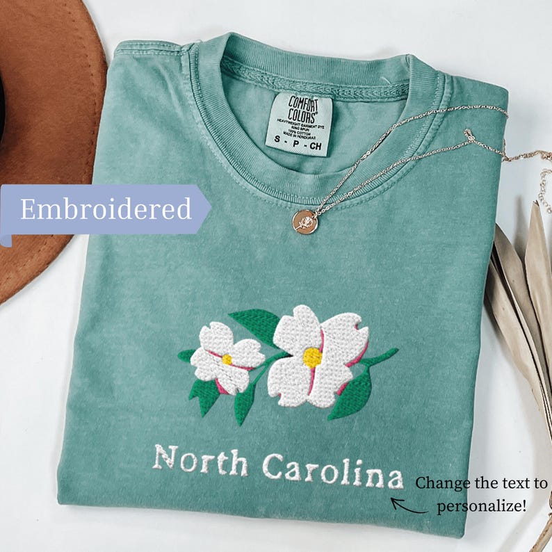 May include: A sage green t-shirt featuring an embroidered design of two white dogwood flowers with yellow centers and green leaves. The text North Carolina is embroidered below the flowers. The shirt has a Comfort Colors label at the neck.