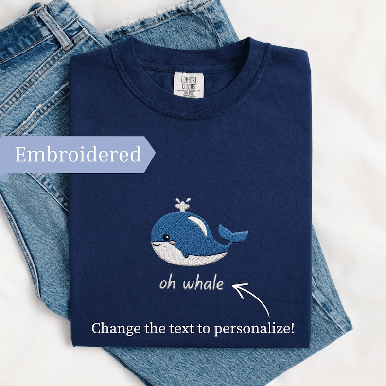 May include: Navy blue t-shirt with an embroidered blue whale design and the text "oh whale". The shirt is folded on top of a pair of blue jeans. The text "Change the text to personalize!" is also visible.