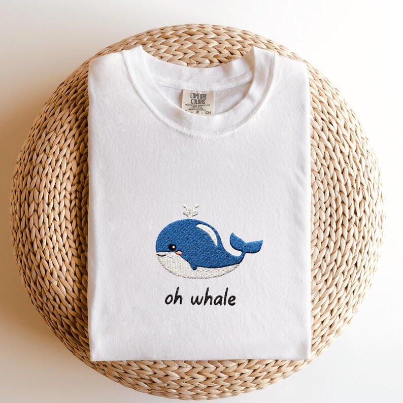 May include: White t-shirt featuring a blue embroidered whale design with the text "oh whale" in black. The whale has a white belly and a small spout. The shirt is made of a soft material.