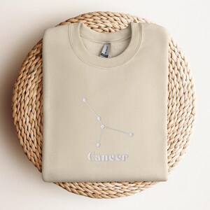 Peut inclure: Un sweat-shirt beige avec une constellation du zodiaque Cancer brod&eacute;e en blanc et le mot "Cancer" en dessous.