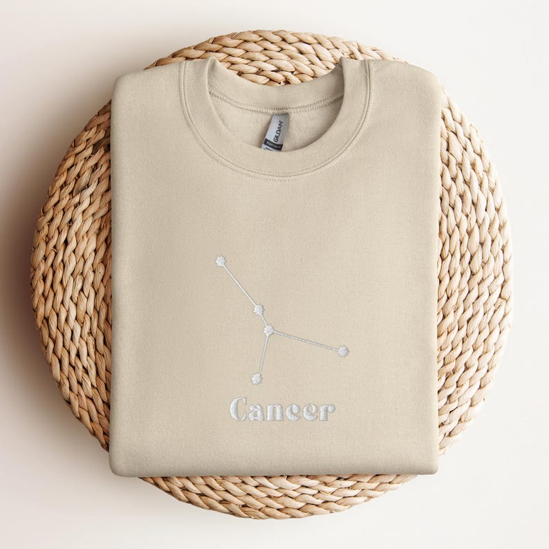 Peut inclure: Un sweat-shirt beige avec une constellation du zodiaque Cancer brod&eacute;e en blanc et le mot "Cancer" en dessous.