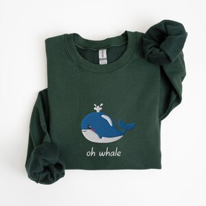 May include: A dark green sweatshirt with a blue whale embroidered on the front. The whale has a white spout and a white belly. The text "oh whale" is embroidered below the whale.