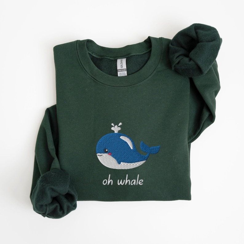 May include: A dark green sweatshirt with a blue whale embroidered on the front. The whale has a white spout and a white belly. The text "oh whale" is embroidered below the whale.