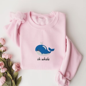 May include: A pink sweatshirt with a blue whale embroidered on the front. The whale has a small white crown on its head and the text "oh whale" is written below it.