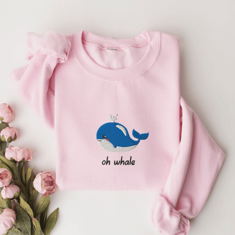 May include: A pink sweatshirt with a blue whale embroidered on the front. The whale has a small white crown on its head and the text "oh whale" is written below it.