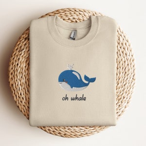 May include: A beige sweatshirt with a blue whale embroidered on the front. The whale has a small crown on its head and the text "oh whale" is written below it.