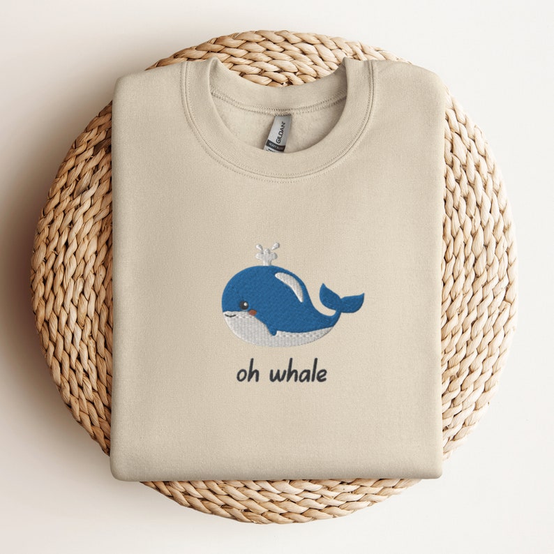 May include: A beige sweatshirt with a blue whale embroidered on the front. The whale has a small crown on its head and the text "oh whale" is written below it.