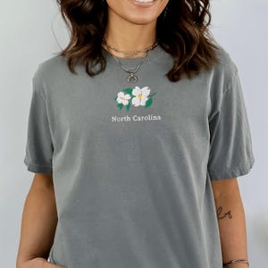 May include: A slate gray short-sleeve t-shirt featuring an embroidered design of two white dogwood flowers with green leaves centered on the chest. Below the floral graphic, the text North Carolina is embroidered in white, sans-serif lettering.