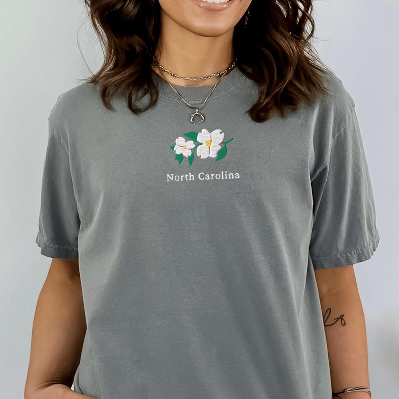 May include: A slate gray short-sleeve t-shirt featuring an embroidered design of two white dogwood flowers with green leaves centered on the chest. Below the floral graphic, the text North Carolina is embroidered in white, sans-serif lettering.