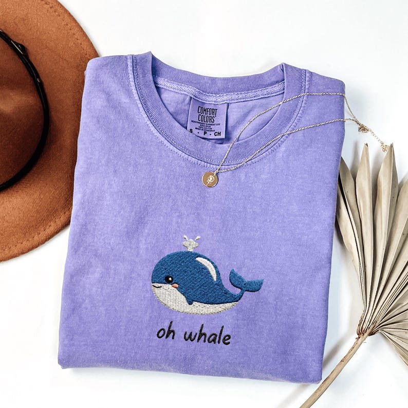 May include: A lavender Comfort Colors t-shirt with an embroidered blue whale design and the text "oh whale". A gold necklace with a pendant is draped across the shirt. The shirt is folded and placed on a white surface.