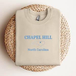 Może przedstawiać: Beżowa bluza z napisem "CHAPEL HILL" i "Karolina P&oacute;łnocna" w niebieskich literach. Stan Karolina P&oacute;łnocna jest zaznaczony na niebiesko z niebieską gwiazdą w środku.