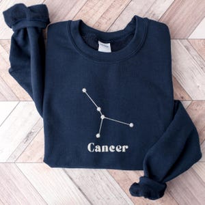 Peut inclure: Un sweat-shirt bleu marine avec une constellation du Cancer brod&eacute;e en blanc et le mot "Cancer" en dessous.