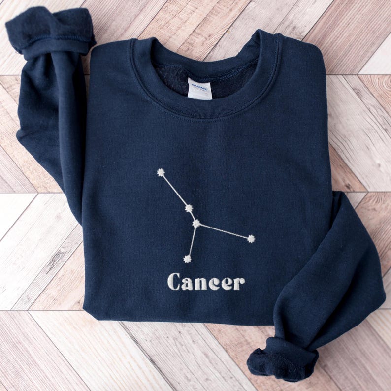 Peut inclure: Un sweat-shirt bleu marine avec une constellation du Cancer brod&eacute;e en blanc et le mot "Cancer" en dessous.