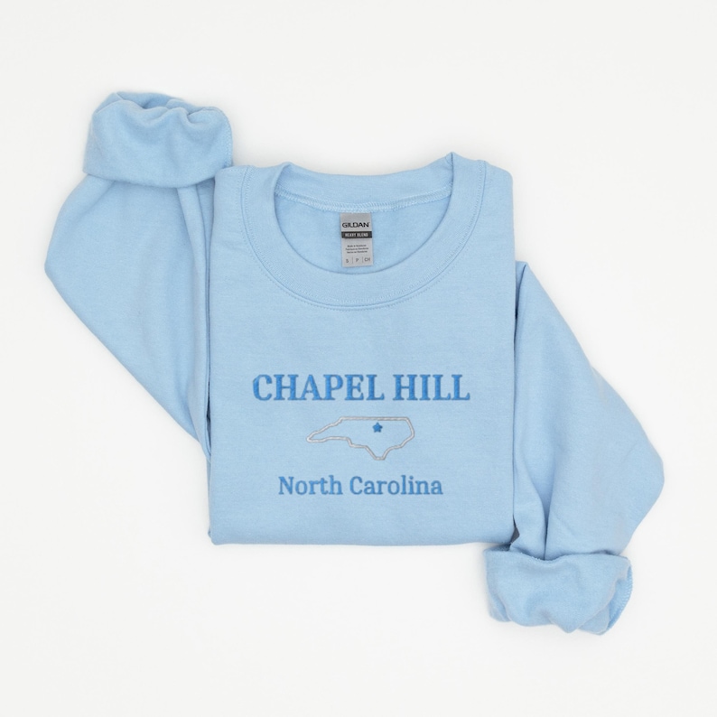 Może przedstawiać: Jasnoszary sweter z napisem "CHAPEL HILL" i zarysowaniem stanu Karolina P&oacute;łnocna z gwiazdą w środku. Napis "North Carolina" znajduje się pod zarysowaniem stanu.