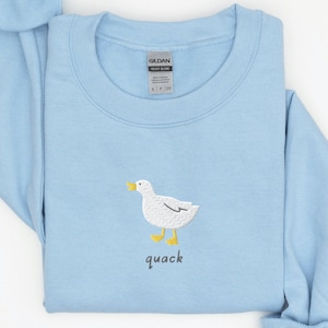以下が含まれることがあります： 薄い青色のスウェットシャツに、白い刺繍の鴨と黒い文字で「quack」と書かれています。