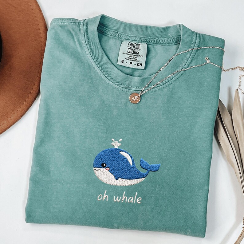 May include: A teal Comfort Colors t-shirt featuring an embroidered blue whale design with the text "oh whale". A silver necklace with a pendant is draped across the shirt. The shirt is a small size.