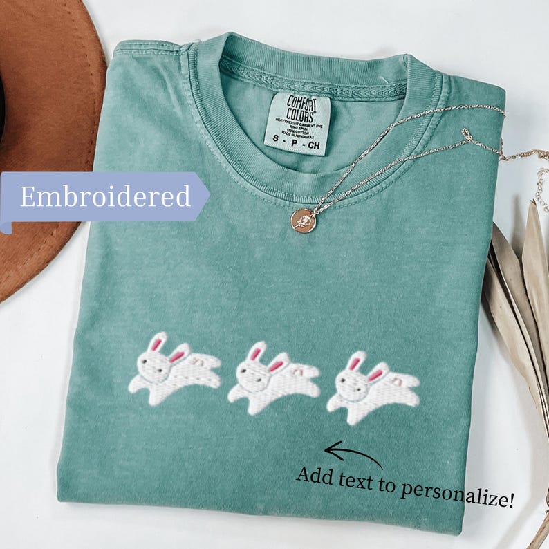 May include: A seafoam green t-shirt with three embroidered white bunnies with pink ears. The shirt has the text "Add text to personalize!" and "Embroidered". A silver necklace with a pendant is on the shirt.
