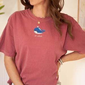 May include: A dusty rose-colored t-shirt featuring an embroidered blue whale design with the text "oh whale" underneath. The shirt has short sleeves and a relaxed fit, perfect for casual wear. The whale is detailed with white accents.