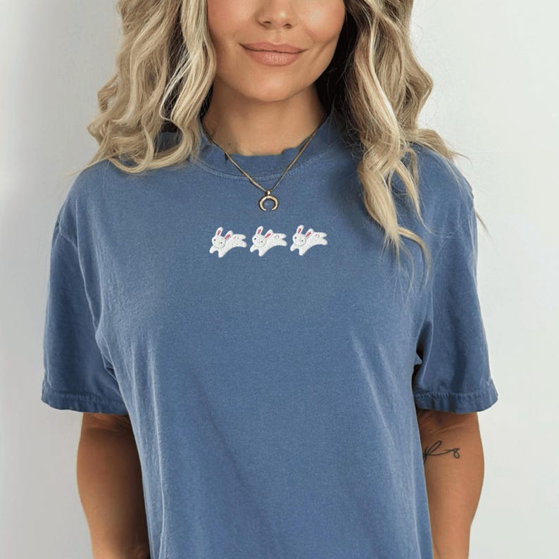 May include: A blue t-shirt with a crew neck and short sleeves. The shirt features three white embroidered bunny rabbits across the chest. The model is wearing a gold necklace.