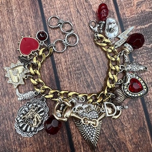 May include: A gold chain bracelet with multiple charms, including a red heart, a horse head, a horseshoe, a sun, a cowboy hat, a bull, and a red crystal heart.