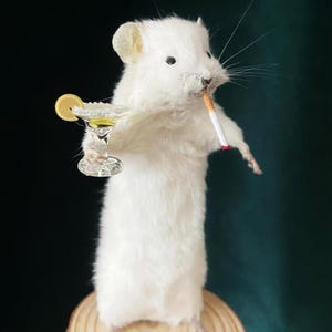 Puede incluir: Un ratón blanco disecado está de pie, sosteniendo un cóctel con una rodaja de limón y un cigarrillo encendido. El ratón está colocado sobre una rodaja de madera sobre un fondo verde oscuro. La copa de cóctel es transparente y adornada.