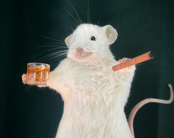May include: A white mouse figurine holding a small glass of amber liquid in one paw and a brown cigar in the other. The mouse has a pink nose and ears, with a long, pink tail. The background is a dark green.