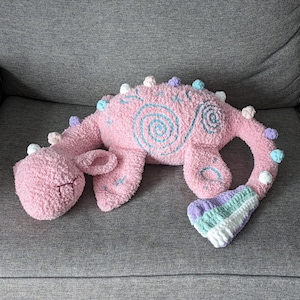 May include: A pink crocheted dragon toy with a blue and white swirl design on its back. The dragon has a long tail with pastel colored pom-poms and a curled up body.