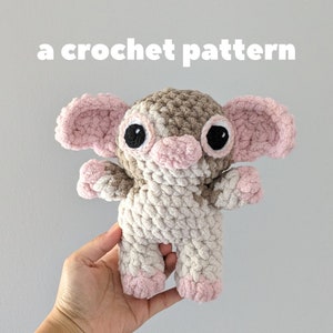 Puede incluir: Un peluche de ganchillo con forma de elefante. El juguete es principalmente de color crema con detalles marrones y rosas en las orejas, la trompa y los pies. Se ven unos grandes ojos negros y el texto "a crochet pattern".