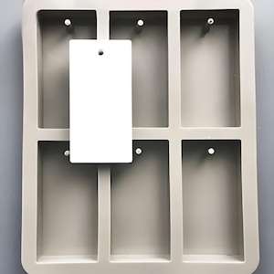 May include: A gray silicone mold with six rectangular cavities. Each cavity has a small hole at the top. A white rectangular tag is placed on top of the mold.