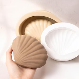 May include: Two silicone molds for making seashell-shaped objects. One mold is white and the other is beige. A brown seashell-shaped object is shown next to the molds.