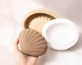 May include: Two silicone molds for making seashell-shaped objects. One mold is white and the other is beige. A brown seashell-shaped object is shown next to the molds.